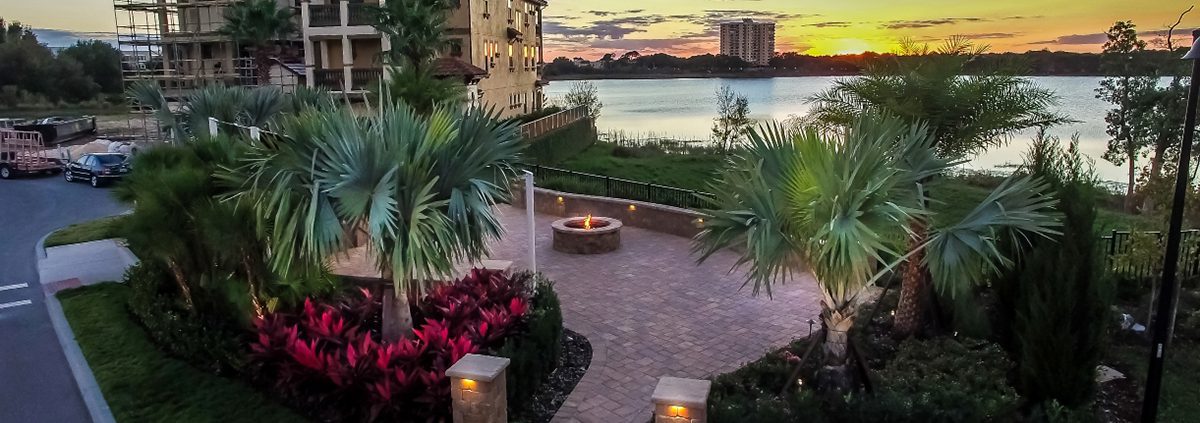 Luxury outdoor living design in Orlando by BLG Environmental, featuring layered palms, tropical plantings, a paver patio, and a lakeside fire pit at sunset.