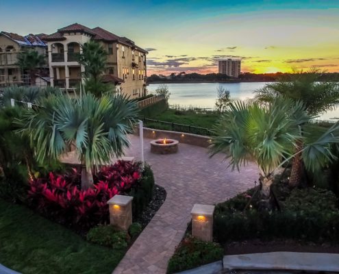 Luxury outdoor living design in Orlando by BLG Environmental, featuring layered palms, tropical plantings, a paver patio, and a lakeside fire pit at sunset.